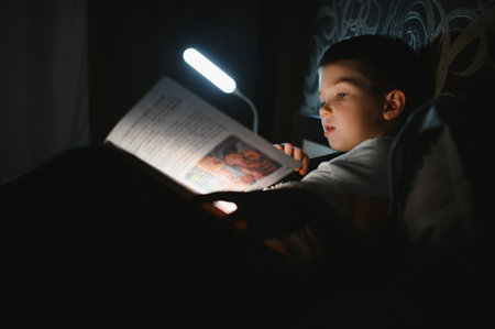 Child reading book in bed. Kids read at night. Little boy with fairy tale books in the bedroom. Education for young children. Bedtime story in the evening. Cute kid under blanket in dark room with lamp.の写真素材