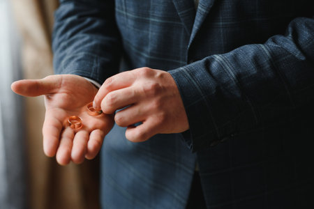 The groom on his wedding day holds two wedding rings in his hand in a hotel room. The man is wearing a white shirt and blue trousers with a vest. Male hand and wedding rings close upの写真素材