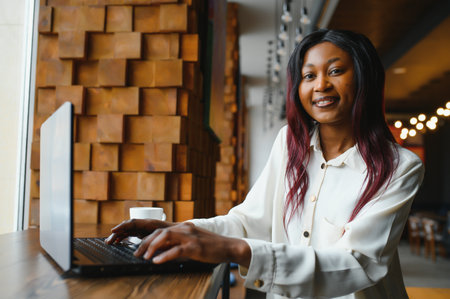 Young African American girl sitting in a restaurant and typing on her laptop. Pretty girl working on computer at cafe.の写真素材