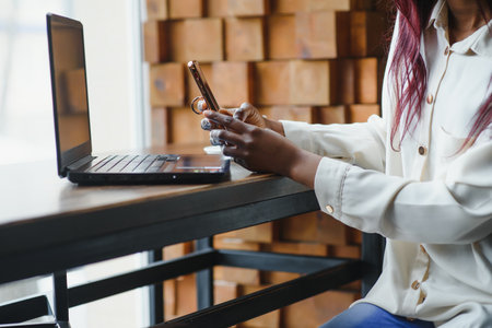 Beautiful african female with a long dark hair is typing messages on a smartphone while sitting beside the window in a soft light. Young business woman is using laptop and mobile phone for the work.の写真素材