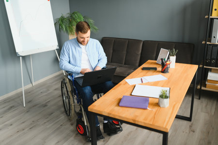 Disabled person in the wheelchair works in the office at the computer. He is smiling and passionate...