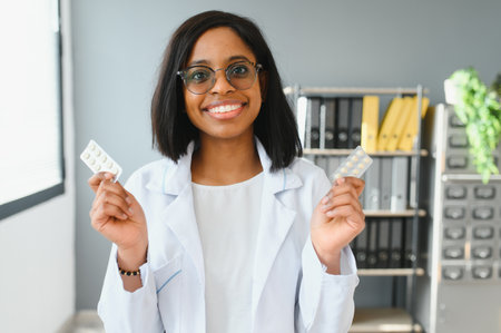 Close up cropped horizontal shot of smiling African female doctor or pharmacist, holding colorful blisters with pills, showing them to camera, while standing in pharmacy or hospitalの写真素材