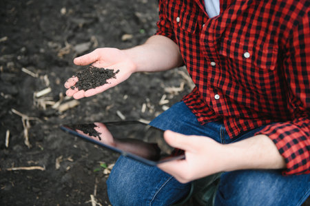 A farmer in boots works with his tablet in a field sown in spring. An agronomist walks the earth, assessing a plowed field in autumn. Agriculture. Smart farming technologies.の写真素材