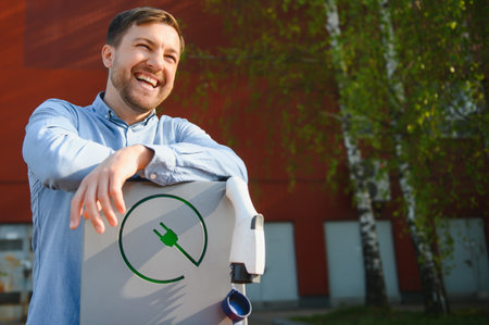 Portrait of a young man standing with charging cable near the charging station. Concept of fast home car chargers.の写真素材