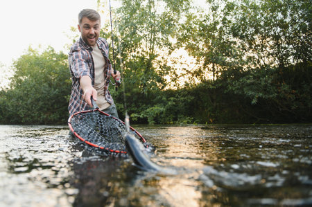 Man with fishing rod, fisherman men in river water outdoor. Catching trout fish in net. Summer fishing hobby.の写真素材