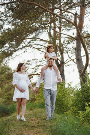 Beautiful pregnant brunette mom and dad and dauther in white clothes look at each other in natureの写真素材