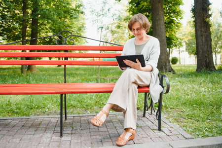 Attractive mature woman sitting on a bench holding and using a digital tablet in the park at summer day.の写真素材
