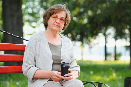 Elegant elderly woman in the shirt is sitting on the bench in a park on a warm day.の写真素材