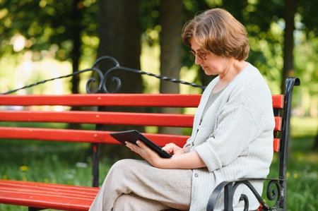 Attractive mature woman sitting on a bench holding and using a digital tablet in the park at summer day.の写真素材