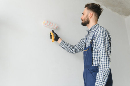 Young smiling professional worker in blue uniform standing with paint roller in new apartment for repairing over gray walls background, copy spaceの写真素材