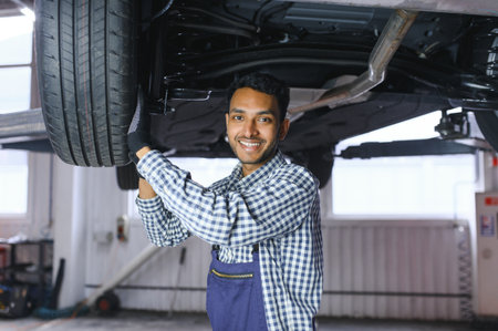 Indian car mechanic standing and working in service station. Car specialists examining the lifted car. Professional repairmen wearing mechanic uniform in blue colorの写真素材