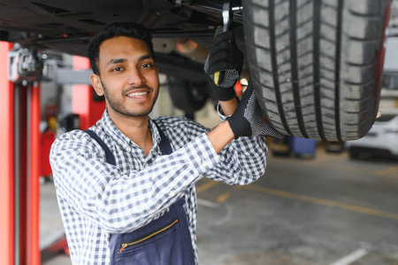 Indian car mechanic standing and working in service station. Car specialists examining the lifted car. Professional repairmen wearing mechanic uniform in blue colorの写真素材