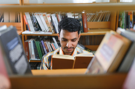 Portrait of cheerful male international Indian student with backpack, learning accessories standing near bookshelves at university library or book store during break between lessons. education concept.の写真素材
