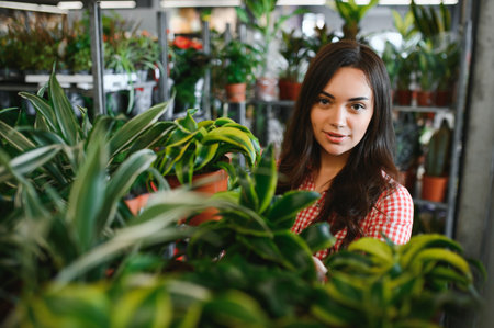 Side rear view of charming female florist in apron putting pots with green plant on shelves in floral shop. Young woman gardener working with houseplants at home. Concept of floral small businessの写真素材