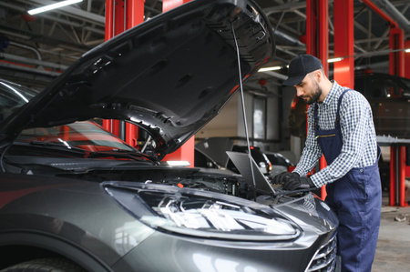 A bearded auto-mechanic in working uniform is standing next to a broken car and using the special laptop for car maintenance. An auto-mechanic in workshop repairs carの写真素材