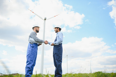 Close up of indian technician and inspector shaking hands while standing on farm with wind turbines. Concept of people, teamwork and eco friendly energy.の写真素材