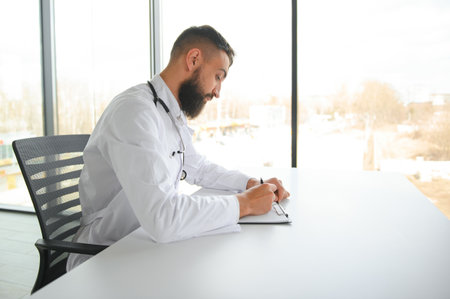 Young 30-aged handsome pleasant Arabic male doctor in white coat, posing at camera indoorsの写真素材
