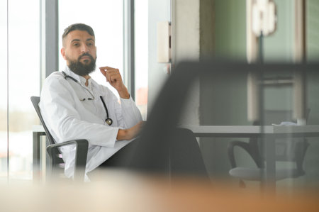 Young 30-aged handsome pleasant Arabic male doctor in white coat, posing at camera indoorsの写真素材