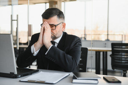Worried businessman in dark suit sitting at office desk full with books and papers being overloaded with workの写真素材