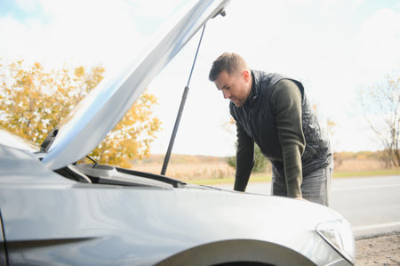 Man repairing a broken car by the road. Man having trouble with his broken car on the highway roadside. Man looking under the car hood. Car breaks down on the autobahn. Roadside assistance conceptの写真素材