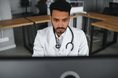 Indian male doctor physician wearing glasses, white medical gown and stethoscope sitting at the desk...