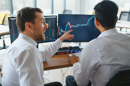 Two men traders sitting at desk at office together monitoring stocks data candle charts on screen...