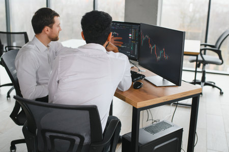 Two men traders sitting at desk at office together monitoring stocks data candle charts on screen...