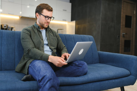 Satisfied handsome young caucasian man in casual stylish clothes, sitting on the couch at home in cozy living room, using a laptop, working or studying online, dreaming, smiling.の写真素材
