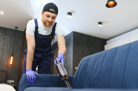 cleaning service. Man janitor in gloves and uniform vacuum clean sofa with professional equipmentの写真素材