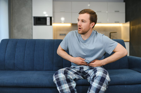 Guy with a beard feeling unwell with a stomach ache while sitting in a couch at home.の写真素材