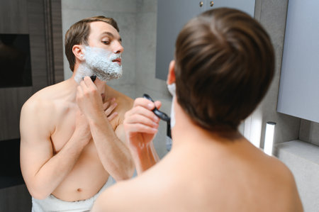 Handsome young man is shaving his face and looking at the mirrorの写真素材