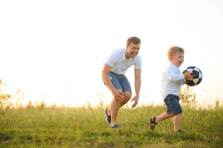 father with a little son plays football on the green grass in the park. Happy family having fun and playing football on a green grassy lawn on a sunny day. Family concept, Father's Day.の写真素材