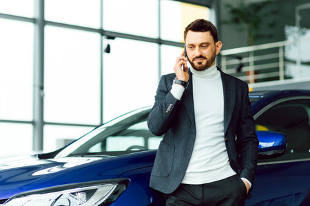 The car shows the character of a man. Confident success elegant business man in full suit and with phone in hand standing near new blue car looks awayの写真素材
