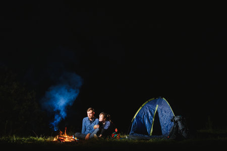 Happy romantic couple travelers resting at bonfire near glowing tourist tent under amazing night sky. The concept of active recreation and travel with a tentの写真素材
