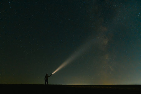 Back view of man with flashlight standing on green grassy field under beautiful dark blue summer starry sky. Night photography, beauty of nature concept. Wide panorama, copy space backgroundの写真素材
