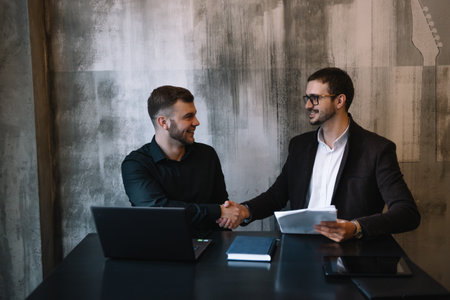Two Businessmen In An Office Smiling At The Camera While Working Together Behind A Laptop Computerの写真素材