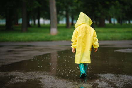 Little boy playing in rainy summer park. Child with umbrella, waterproof coat and boots jumping in puddle and mud in the rain. Kid walking in summer rain Outdoor fun by any weather. happy childhoodの写真素材