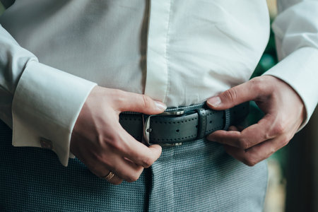 A man in blue pants and a white shirt buttoned a brown leather trouser belt. He has a watch on his...
