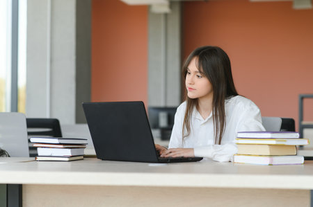 A beautiful caucasian female student is studying in college remotely. She is sitting with a laptop and a notepad and concentrated is watching a video conference lesson.の写真素材