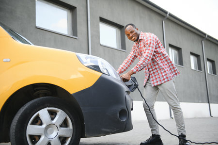 A african american man stands next to yellow electric delivery van at electric vehicle charging stations.の写真素材