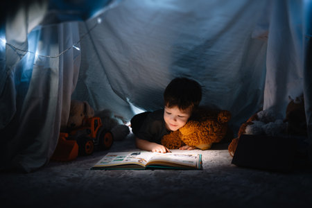 child boy reading with book and flashlight and teddy bear in tent. before going to bedの写真素材