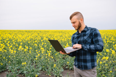 Young agronomist holds laptop in rape field. Agribusiness concept. agricultural engineer standing in a rape field with a laptop in summerの写真素材