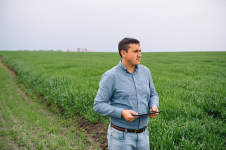 young handsome agronomist, agriculture engineer standing in green wheat field with tablet in hands...