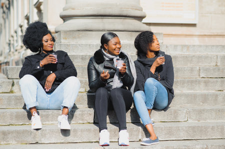 three beautiful and stylish dark-skinned girls with long hair standing in a city and drinking a coffee and use the phonesの写真素材