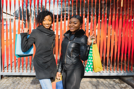 Portrait of young two african ladies friends standing over red wall and posing with shopping bags.の写真素材