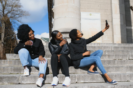 three beautiful and stylish dark-skinned girls with long hair standing in a city and drinking a...