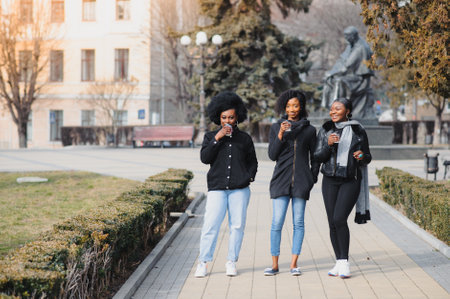 three beautiful and stylish dark-skinned girls with long hair standing in a city and drinking a coffee and use the phonesの写真素材