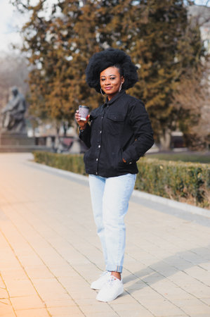 Cheerful dark skinned woman enjoying coffee holding to go cup recreating in city park, happy trendy dressed african american hipster girl sitting outdoors during sunny day drinking beverageの写真素材