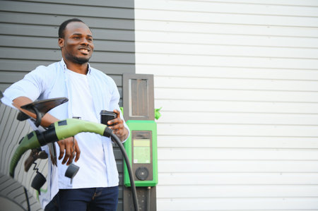 Stylish african man with coffe cup in hand inserts plug into the electric car charging socket.