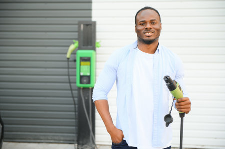 Serious african man holding charge cable in on hand, standing near luxury electric carの写真素材